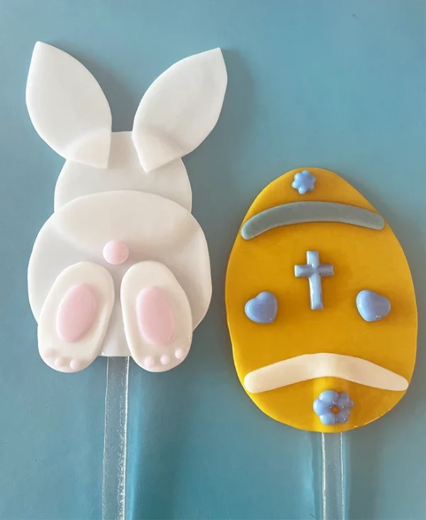 A pair of whimsical easter-themed garden yard steak decorations, with one resembling a bunny from behind and the other shaped like an egg with a cross and decorative elements made at the Glass Duchess Studio in Port Charlotte, FL